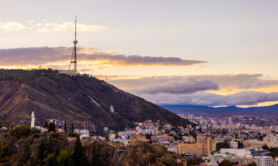 Tbilisi showcases a vibrant skyline during sunset, with the TV tower prominently displayed on a mountain, while clouds add depth to the evening sky.