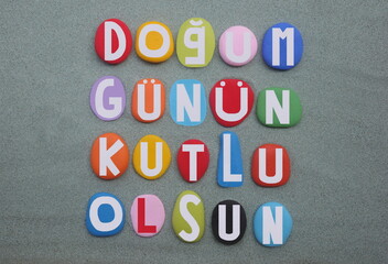 Dogum gunun kutlu olsun, Happy birthday in turkish language composed with multi colored hand painted stone letters over green sand for a unique postcard, banner or poster 