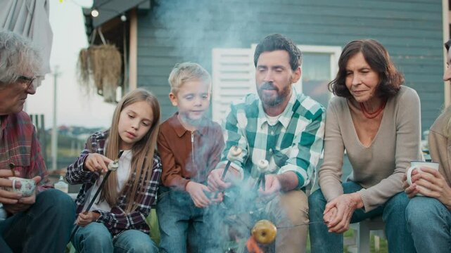 Grandchildren visiting their grandparents in village. Family sitting around campfire. Preparing delicious marshmallow and fruits. Kids taking hot dessert from wooden sticks. Spending time in nature.