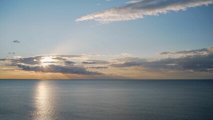 Fototapeta premium Sun shining through clouds over calm ocean at sunset