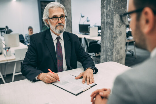 Successful formal dressed senior businessman with pen in hands sitting at meeting table and discussing business plan and organization of working process during brainstorming in financial company.