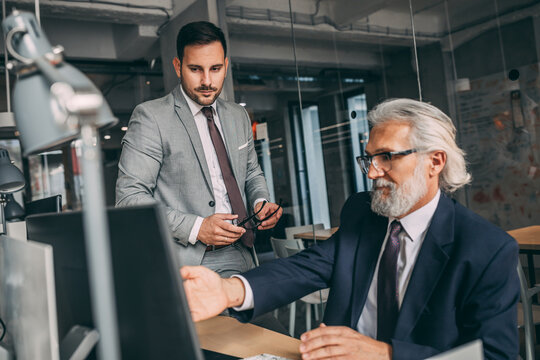 Business people sitting at desk in the modern office, looking at PC screen and discussing project, confident mature man explaining something to his young male coworker
