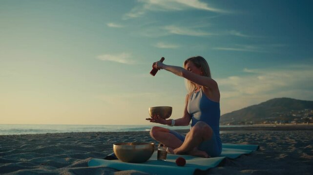 A woman sits on a yoga mat at the beach during sunset, playing Tibetan singing bowls for sound healing and meditation. Peaceful scene.