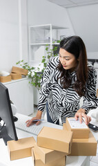 Young Asian inventory manager is preparing a package to ship, working on a computer to check order...