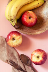 Bananas and  red apples laying on a wooden plate seen from above. Wooden fork and spoon  alongside a dish towel. Pink background . Natural light, bright and colorful.