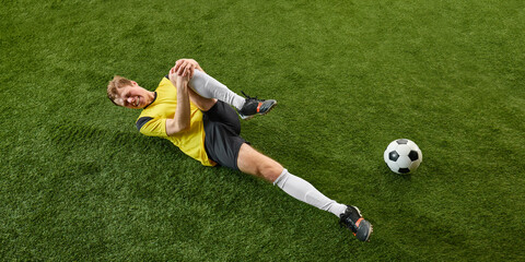 Young man, soccer player lying on stadium with injured leg, expressing emotions of pain. Intense play. Concept of sport, action, competition, game, active lifestyle. Ads