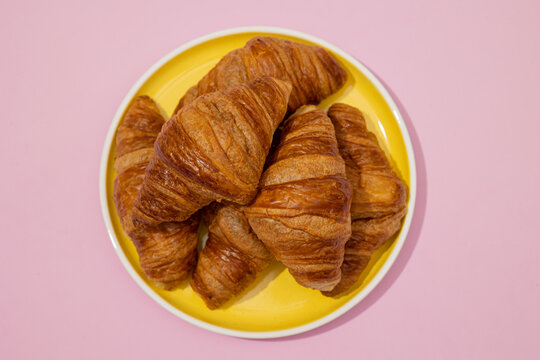 Top view of croissants piled on a yellow plate on a minimalist pastel pink background, puff pastry bakery piece