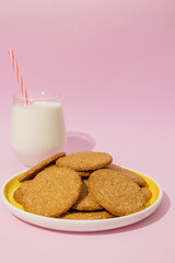 Vertical image of whole wheat oatmeal cookies on a yellow plate with a glass of milk next to it on a pastel pink background, cookies made from wheat flour mixed with oat cereal, traditional in baking