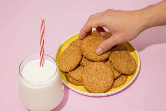 Male hand taking whole grain oatmeal cookies from a yellow plate with a glass of milk next to it on a pastel pink background, cookies made from wheat flour mixed with oat cereal, traditional in baking