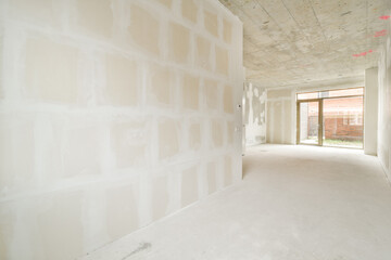 A spacious interior in the process of construction, featuring bare walls and a concrete ceiling. Natural light filters through an opening, highlighting the unfinished space.