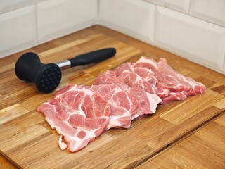 Slices of succulent pork ready for seasoning on a rustic wooden kitchen countertop with a meat mallet nearby