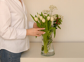 Close-up of a woman’s hands placing fresh white tulips in a glass vase. The minimalistic and cozy setting creates a soft and elegant atmosphere, perfect for home decor and lifestyle concepts.