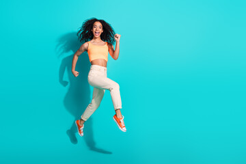Energetic young woman jumping joyfully in vibrant summer outfit against a vivid teal background,...