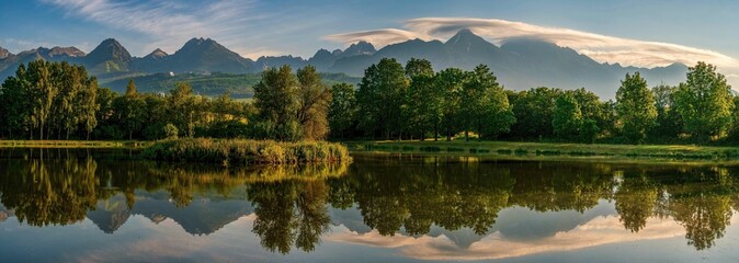 Fototapeta premium Summer mountain landscape with lake, trees and peaks in the background, reflection of mountains and landscape in the lake. Strba Lake under the Peaks of High Tatras Mountains Lit by Golden Ligh