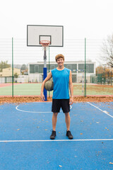Fototapeta premium Portrait of a male basketball player looking at camera holding the ball under his arm in a basketball court. Male in sportswear standing on outside basketball court with ball ready to play game.