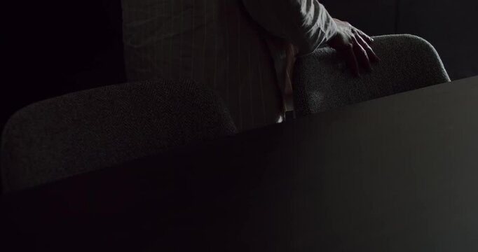 A person rests their hand on a chair in a dimly lit, moody office environment. Minimalist design with dark tones. Person Leaning on Chair in Dark Moody Office Setting, Minimalist Design