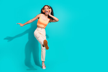 Energetic young woman enjoying music while dancing against a teal background, dressed in a vibrant outfit