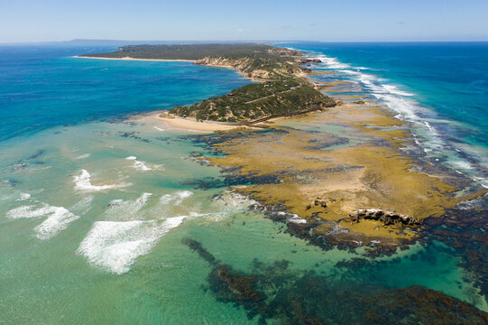 An aerial view of waves crashing onto a rocky platform at the end a a long coastal peninsula