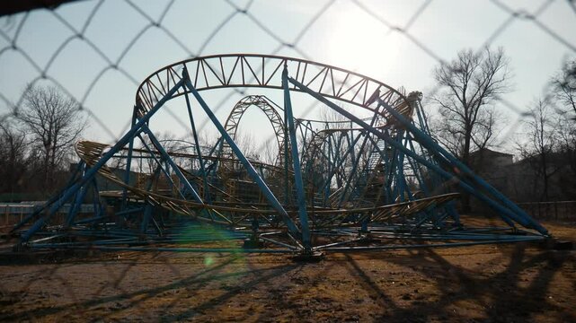 Video shooting through chain link fence rust non-working roller coaster in abandoned amusement park on sunny day.