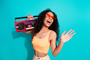 Joyful young woman holding a vintage red boombox, wearing stylish orange sunglasses and top,...
