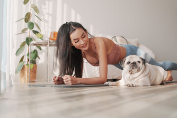 Beautiful Asian woman performs a plank exercise with her pug beside her in a living space,...