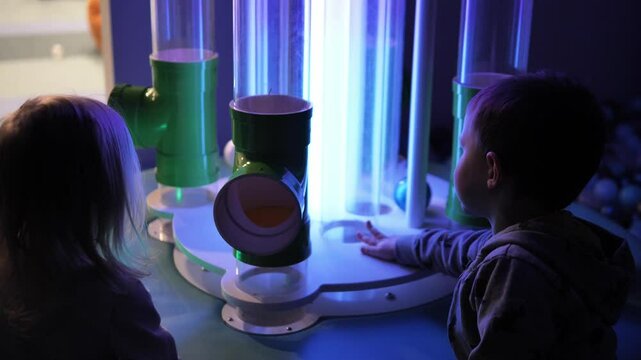Two young children, a boy and a girl, explore a fascinating interactive exhibit with glowing tubes and balls, their curiosity piqued by the mesmerizing display of light and motion