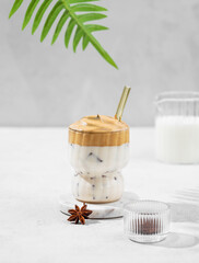 Dalgona coffee in a glass on a light background with milk jug, spices and palm leaf.