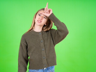 beautiful teen girl wearing green sweater making fun of people with fingers on forehead doing loser...