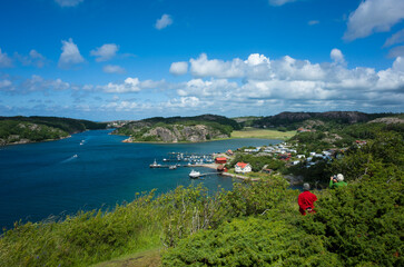 Scenic coastal landscape south of Hamburgsund, Sweden. A blue bay with boats, rocky shores, and a camping. An elderly couple stands on a green hillside, admiring the summer view under a cloudy sky