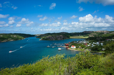 Scenic coastal landscape on Sweden’s west coast, south of Hamburgsund. A vibrant blue bay with boats, green hills, rocky shores, and a camping site near the shoreline under a partly cloudy summer sky