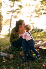 Mom sits on a stump and kisses on the cheek a little girl standing next to her in the forest