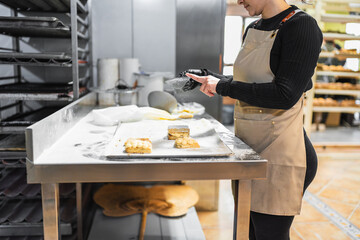 Pastry chef sprinkling icing sugar on delicious pastries in bakery kitchen