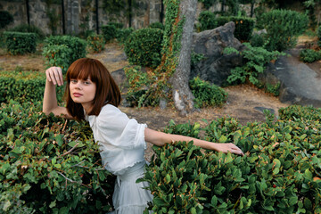 Elegant woman in white dress leaning against lush greenery in serene garden setting