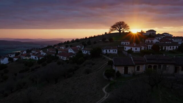 Orange and purple sunset over a small village with a prominent tree silhouetted against the setting sun