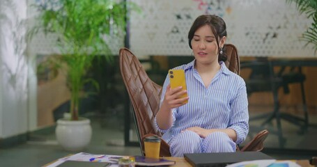Afghan young adult gen z woman lady sitting on chair use mobile look cam talk gossip indoor job place happy girl staff hold phone speak on video call after work enjoy break spend day time at office