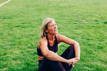 Tired middle-aged woman resting after a workout - Athletic middle-aged woman in sportswear sitting on the grass, catching her breath after exercising outdoors in a park