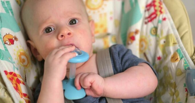 baby in blue holding and chewing on a teething toy, lies on a soft play mat, infant looks curious and engaged, exploring textures while soothing sore gums, home comfort, teething relief, baby care.