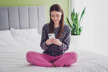 Young woman sitting on bed in pajamas using smartphone during a relaxed morning indoors with...