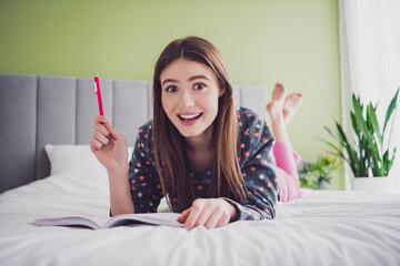 Young woman lying on bed in pajamas, smiling with enthusiasm, holding a pen, enjoying a relaxed and...