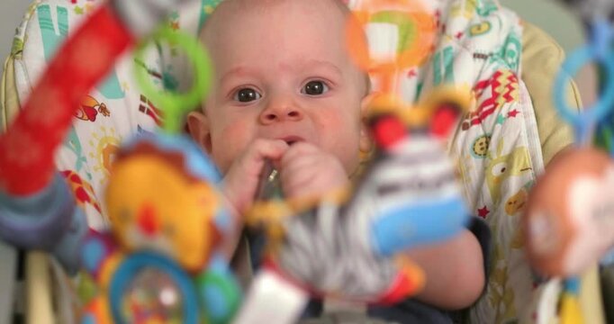  baby sits in baby seat, surrounded by colorful hanging toys, infant looks at objects curiously while bringing its hand to the mouth, engaging in sensory exploration, early learning, playtime
