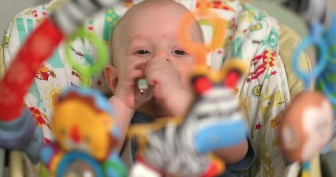  baby sits in baby seat, surrounded by colorful hanging toys, infant looks at objects curiously while bringing its hand to the mouth, engaging in sensory exploration, early learning, playtime