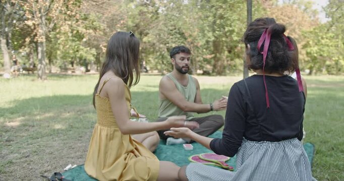 Friends enjoying a summer picnic in the park, playing a hand-clapping game and sharing laughter together.