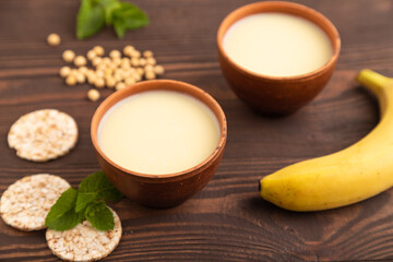 Organic non dairy banana and soy milk in clay cup on brown wooden, side view, selective focus.