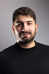 Obraz premium portrait d'un beau jeune homme de 25 ans. Il est brun, barbu et souriant et il porte un pull over noir. Arrière plan gris