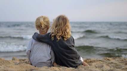 Blonde sisters sitting closely on sandy shore, embracing while gazing at ocean waves during...
