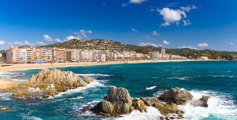 Vista panorámica de la Playa de Lloret en Costa Brava, con mar azul, rocas, arena dorada y edificios frente al mar. © jocaja