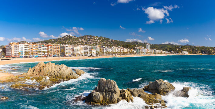 Vista panor&aacute;mica de la Playa de Lloret en Costa Brava, con mar azul, rocas, arena dorada y edificios frente al mar.