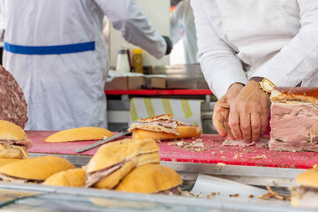 Vendor in a white coat prepares panini with sliced prosciutto on a red cutting board while assembling sandwiches with fresh bread and meat at an Italian market stall