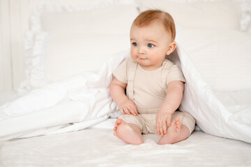 Fototapeta premium A baby on a white bed under a blanket is smiling, a cute little baby boy with blue eyes in beige clothes woke up on a white bed in the bedroom at home.