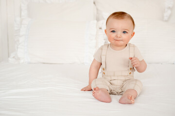 Fototapeta premium A baby on a white bed is smiling, a cute little baby boy in beige clothes is sitting on the bed in the bedroom at home, a smiling baby